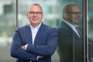 Bewerbungsfotos im Rostocker Fotostudio: Geschäftsmann im blauen Anzug mit Brille steht selbstbewusst mit verschränkten Armen vor einer Glasfassade. Sein Spiegelbild ist im Hintergrund sichtbar.