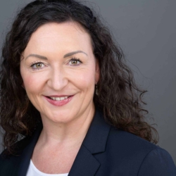 Bewerbungsfotos im Rostocker Fotostudio – Professionelles Business-Portrait einer Frau mit lockigen dunklen Haaren, freundlichem Lächeln und dunklem Blazer vor grauem Hintergrund.