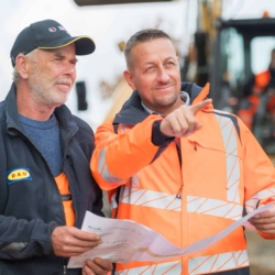 Zwei Bauarbeiter in Sicherheitskleidung besprechen Baupläne auf einer Baustelle, im Hintergrund ein Bagger – fotografiert vom Businessfotograf in Rostock.