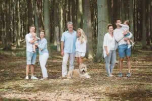 Drei Generationen einer Familie stehen lachend zusammen im lichten Wald, inklusive zwei Kleinkindern und einem freundlichen Hund im Vordergrund. Alle tragen helle, sommerliche Kleidung. Foto von Martin Börner, Familienfotograf aus Rostock.
