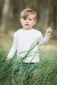 Kleiner Junge mit blonden Haaren und weißem Pullover steht im hohen grünen Gras und schaut neugierig in die Kamera.
