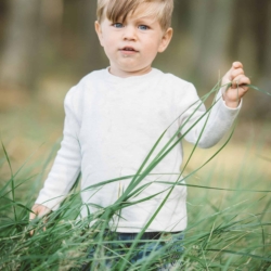 Kleiner Junge mit blonden Haaren und weißem Pullover steht im hohen grünen Gras und schaut neugierig in die Kamera.