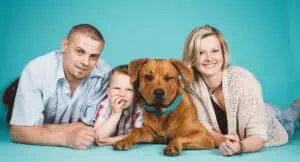 Familienfoto im Fotostudio: Eltern und ihr kleiner Sohn liegen mit ihrem Hund auf dem Boden vor einem blauen Hintergrund und strahlen Freude aus.