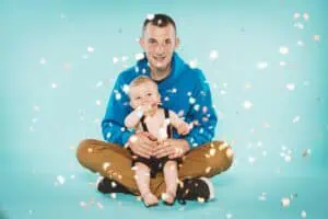 Vater und Baby sitzen gemeinsam auf dem Boden, umgeben von buntem Konfetti – fröhliches und farbenfrohes Fotostudio-Portrait.