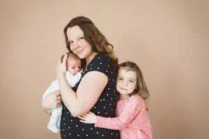 Mutter mit Neugeborenem auf dem Arm und Kleinkind, das sie von hinten umarmt – liebevolles Familienportrait, aufgenommen vom Fotografen zu Hause.