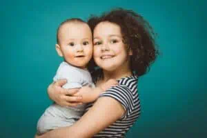 Lächelndes Geschwisterpaar im Fotostudio vor türkisfarbenem Hintergrund – älteres Mädchen hält Babybruder im Arm, fröhliches und professionelles Geschwisterfoto.