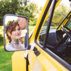 Braut mit Blumenkranz und Tattoo lächelt in die Kamera, während der Bräutigam sie auf die Stirn küsst – festgehalten im Rückspiegel eines gelben Fahrzeugs.