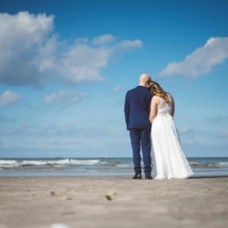 Brautpaar in Hochzeitskleidung steht Hand in Hand am Ostseestrand in Warnemünde und blickt gemeinsam aufs Meer bei blauem Himmel.