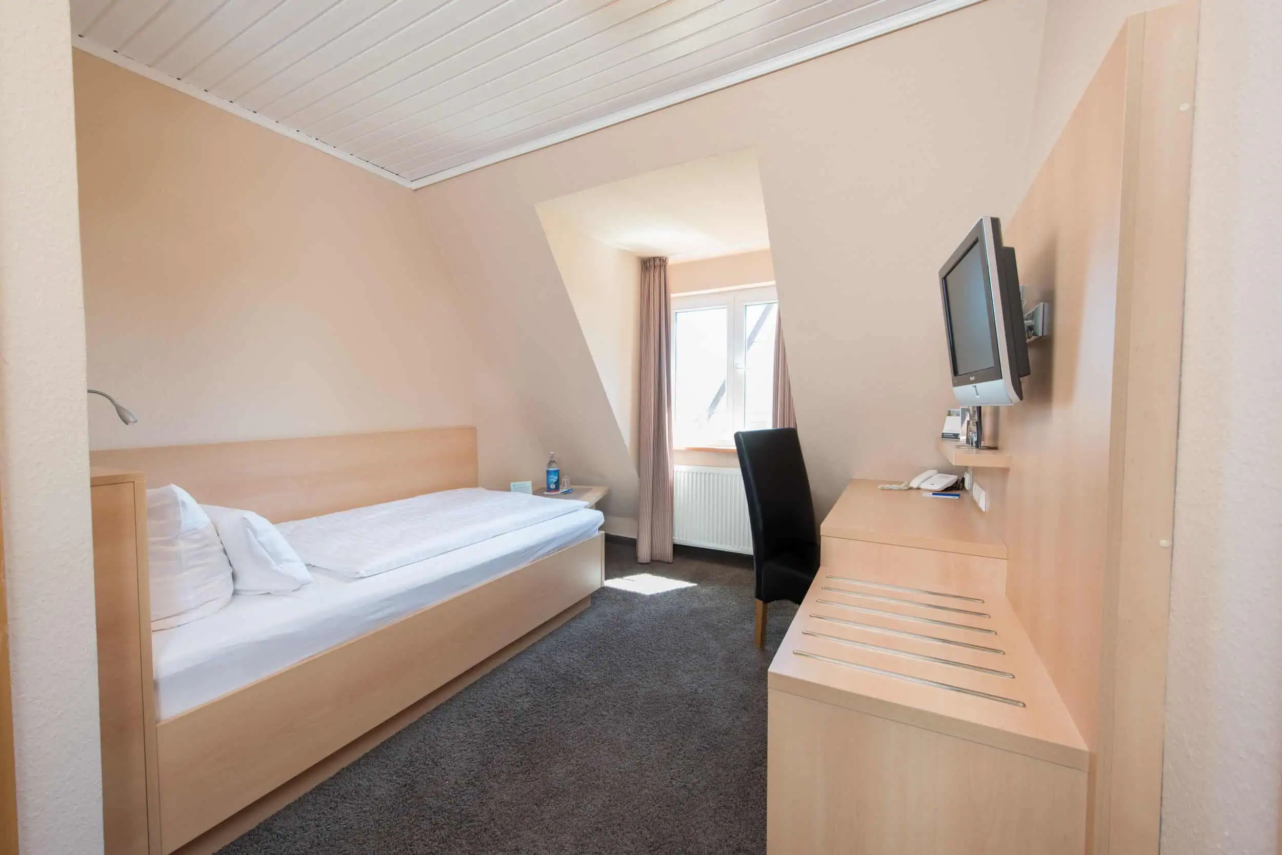 hotel-am-alten-strom-zimmer-interior-fotograf-003 Einzelzimmer im Hotel Am Alten Strom mit hellem Holzmobiliar, Schreibtisch, Fernseher und Fenster mit Tageslicht.