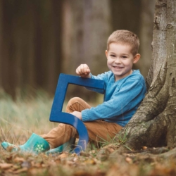 Junge sitzt lachend mit einer großen blauen Zahl Fünf an einem Baum im herbstlichen Wald.