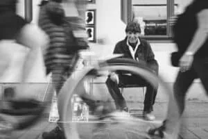 Portraitfotograf Rostock – Schwarz-Weiß-Porträt eines Mannes auf einem Stuhl, umgeben von vorbeigehenden Passanten und Fahrrädern.