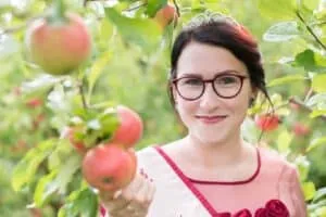 Outdoor Porträt einer jungen Frau mit Krone und Brille, die einen Apfel in einem blühenden Obstgarten in der Hand hält.
