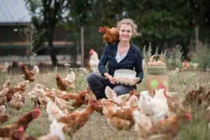 Junge Frau sitzt auf einer Wiese zwischen vielen braunen Hühnern, hält eine Schachtel frischer Eier in der Hand, während ein Huhn auf ihrer Schulter sitzt. Im Hintergrund sind Eierkartons mit der Aufschrift „Eier Abo“ zu sehen. Foto von Martin Börner, Rostock.