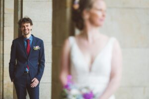 Bräutigam im Fokus beim kreativen Hochzeitsshooting in Rostock Bräutigam in dunkelblauem Anzug mit roter Krawatte lächelt glücklich, während die Braut im weißen Kleid unscharf im Vordergrund steht – modernes