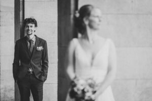 Kreatives Brautpaarfoto mit Fokus auf den Bräutigam – Hochzeit in Rostock Der Bräutigam steht lächelnd an einer hellen Steinwand, während die unscharf im Vordergrund stehende Braut im weißen Kleid mit Blumenstrauß abgewandt blickt – kreative Hochzeitsfotografie in Rostock.