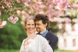 Romantisches Brautpaarshooting unter Blüten in Rostock Lächelnde Braut in weißen Hochzeitskleid wird von ihrem Bräutigam liebevoll von hinten umarmt, umgeben von rosa Kirschblüten während des Brautpaarshootings in Rostock.