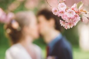 Kirschblüten im Fokus mit Brautpaar im Hintergrund in Rostock Zarter Zweig mit rosa Kirschblüten im Fokus, dahinter unscharfes küssendes Brautpaar beim Hochzeitsshooting in Rostock.