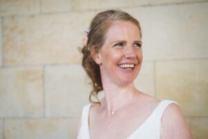 Portrait einer fröhlichen Braut in natürlichem Licht – Hochzeitsfoto mit Herz in Rostock Strahlende Braut mit offenem Lächeln vor heller Natursteinwand – natürliche Hochzeitsfotografie mit emotionalem Charme.