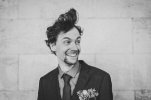 Lachender Bräutigam mit wilder Frisur – authentisches Hochzeitsfoto mit Humor Bräutigam mit lockerem Look und fröhlichem Lachen vor heller Steinwand – seine Frisur steht wild nach oben.