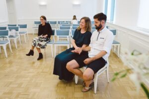 Hochzeitsfotos im Standesamt Rostock – echte Momente der Gäste in sommerlicher Stimmung Hochzeitsgäste im Standesamt Rostock in sommerlicher Kleidung – eine lachende Frau hält die Hand ihres Partners, umgeben von leeren Stühlen.