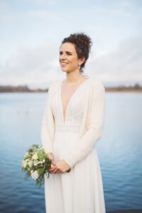 Natürliches Brautportrait mit Cardigan am Stadthafen Rostock Braut mit weißem Strick-Cardigan und Blumenstrauß posiert lächelnd vor dem Wasser am Stadthafen Rostock.