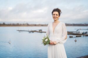 Zeitloses Brautportrait am Wasser – Hochzeitsshooting am Stadthafen Rostock Braut in elegantem, fließendem Hochzeitskleid mit Blumenstrauß posiert vor dem Wasser des Stadthafens Rostock bei bewölktem Himmel.