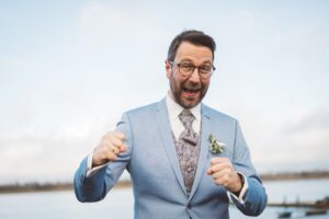 Bräutigam Portrait mit Energie und Stil am Stadthafen Rostock Fröhliches Portrait eines Bräutigams im blauen Anzug mit floraler Weste am Stadthafen Rostock, mit erhobenen Fäusten und breitem Lächeln.