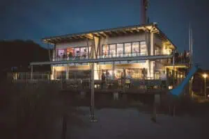 Abendaufnahme der Hochzeitslocation Blaue Boje in Markgrafenheide mit beleuchtetem Gebäude und Gästen auf der Terrasse.