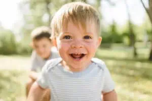 Lachender kleiner Junge mit blonden Haaren in einem gestreiften Shirt im Sonnenschein, unscharfer Junge im Hintergrund im Park.