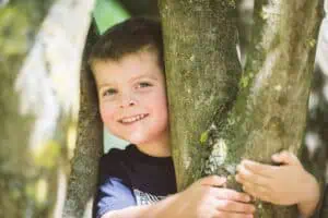 Lächelnder Junge lehnt sich an einen Baumstamm und schaut in die Kamera – Kinderportrait im Freien.