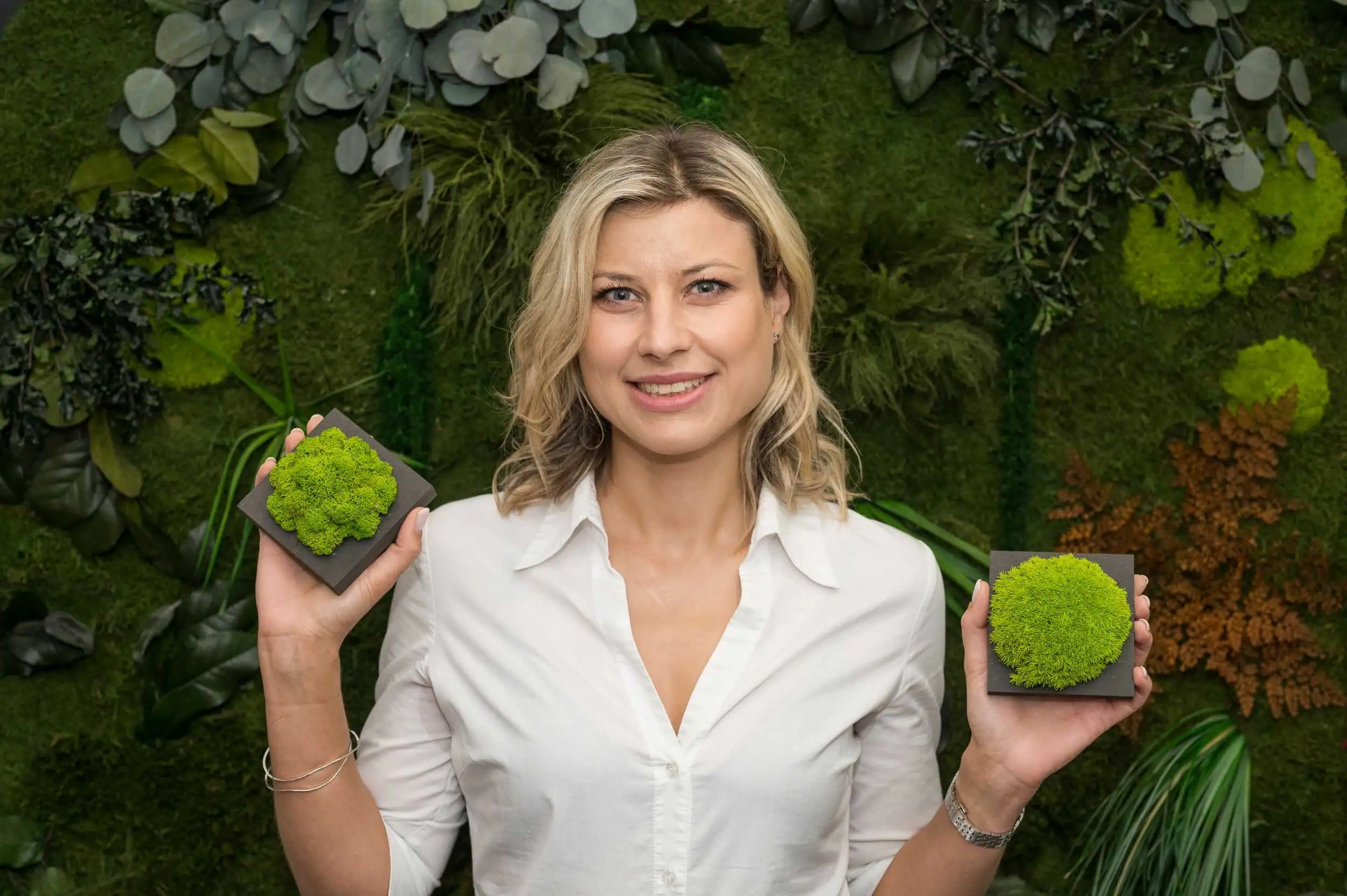 Moos Antje Gude präsentiert zwei Moosproben vor einer grünen Pflanzenwand, umgeben von verschiedenen Blattstrukturen und Naturmaterialien.