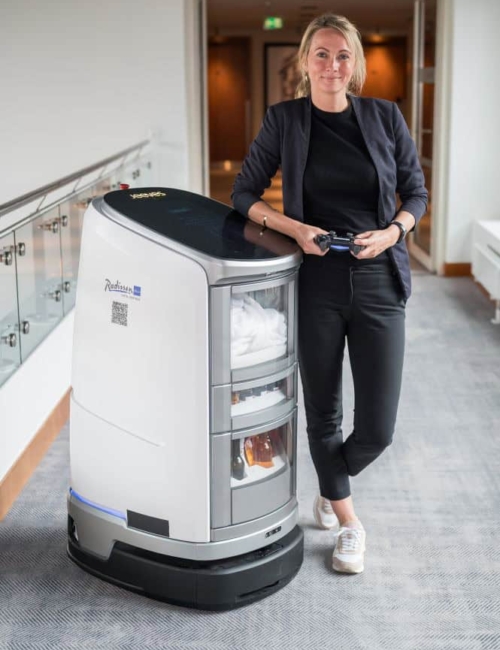 Roboter Jeeves im Radisson Blu Rostock Mitarbeiterin im Radisson Blu Hotel steht mit Fernbedienung neben einem modernen Hausroboter mit Handtüchern und Pflegeprodukten.