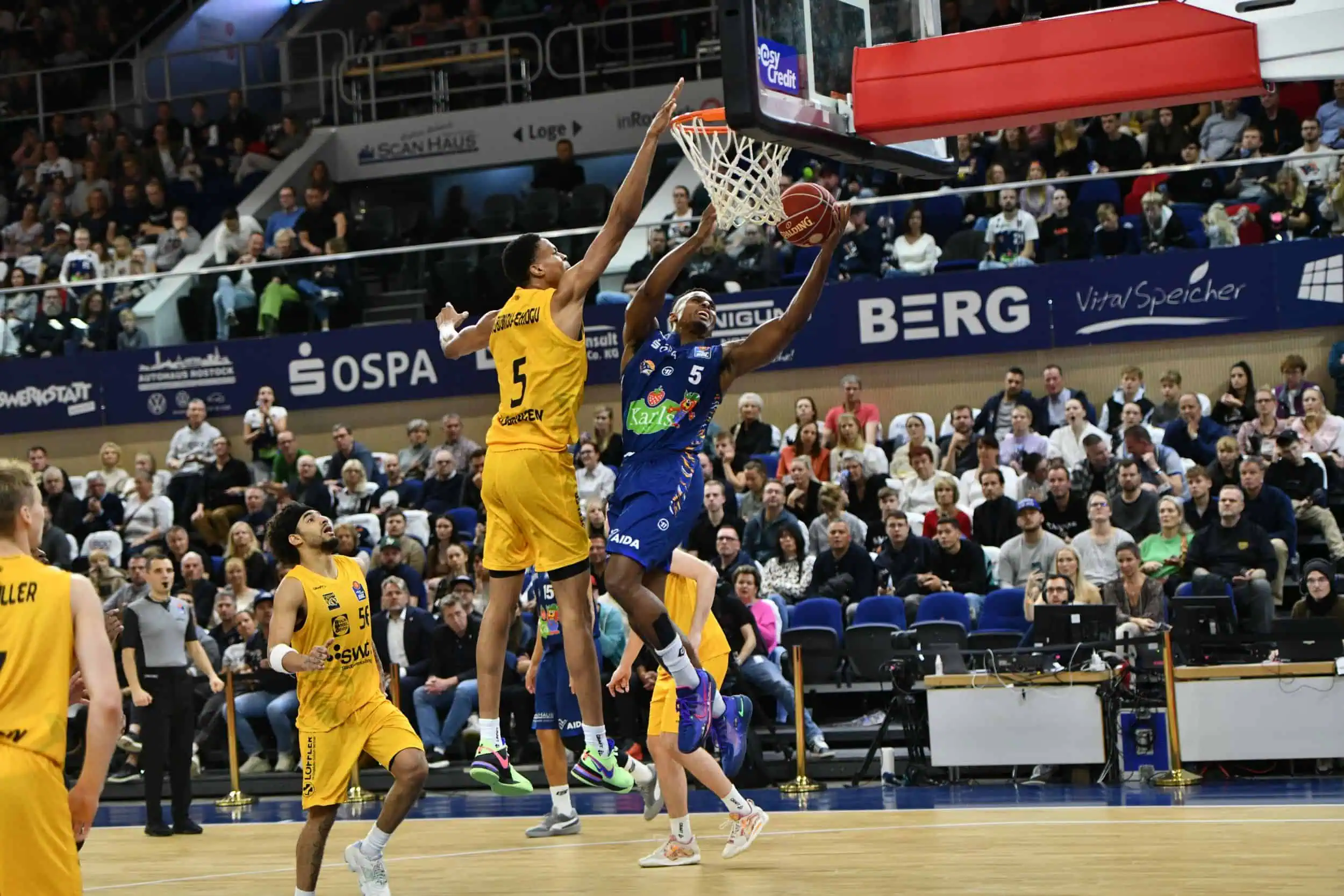 event-basketballspiel-rostocker-seawolves-tübingen-stadthalle-029 Spieler der Rostock Seawolves im blauen Trikot beim Korbleger gegen gelb gekleidete Gegner in ausverkaufter Arena.