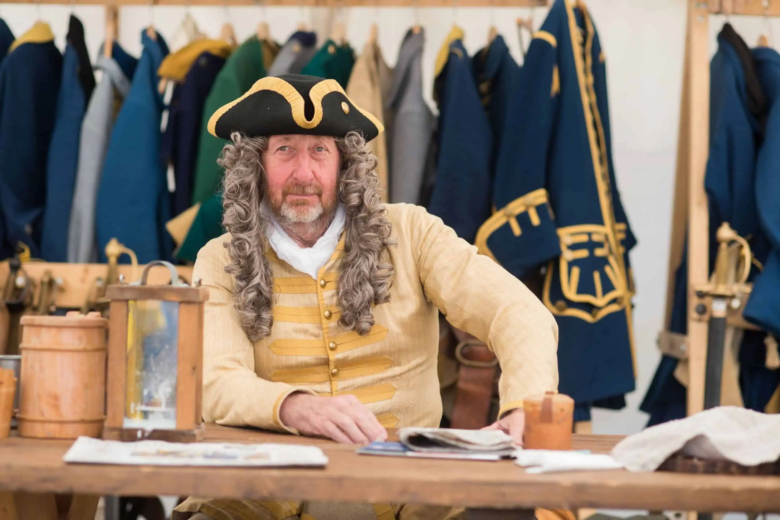 event-schwedenfest-wismar-foto-schweden-006 Darsteller mit Perücke und Uniform sitzt am Holztisch beim Schwedenfest Wismar vor historischen Kostümen.
