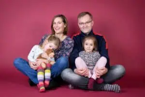 Fröhliche Familie im Fotostudio: Mutter, Vater und zwei Töchter sitzen auf dem Boden vor einem rosa Hintergrund. Ein Mädchen hält ein Kuscheltier, alle lachen und strahlen.