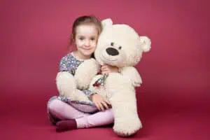 Lächelndes Mädchen sitzt vor rosa Hintergrund im Fotostudio und umarmt einen großen Teddybären – charmantes Kinderporträt.