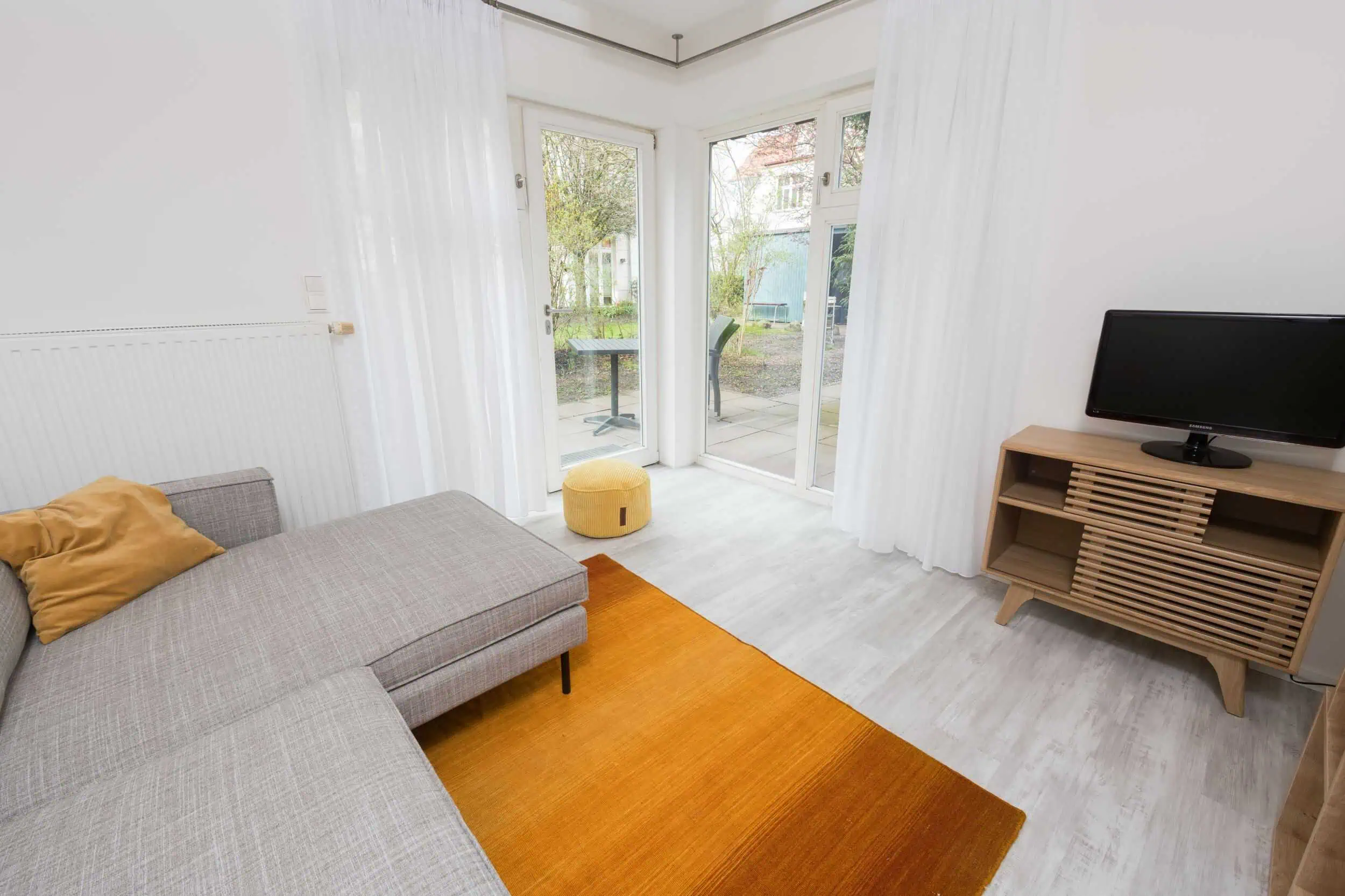 fotograf-rostock-ferienwohnung-innenaufnahmen-018 Heller Wohnbereich einer Ferienwohnung mit Terrasse in Kühlungsborn, ausgestattet mit grauem Sofa, Fernseher, gelbem Teppich und Zugang zur möblierten Außenterrasse.