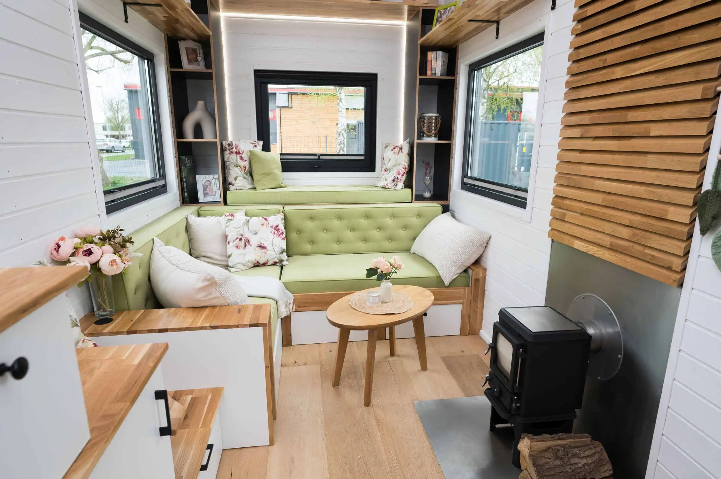 So lebt es sich im Tiny House Gemütlicher Wohnbereich in einem modern eingerichteten Tiny House von Mobi Haus mit grünem Sofa, Holzboden, Holzofen und dekorativen Elementen.