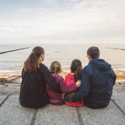 Familie mit zwei Kindern sitzt Rücken an Rücken auf einer Promenade am Meer und blickt gemeinsam in die Ferne über das Wasser.