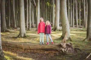 Zwei Mädchen stehen auf einem umgefallenen Baumstamm im Wald und blicken in die Kamera.