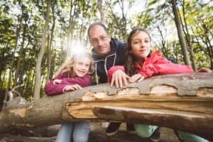 Vater mit zwei Töchtern lehnt lachend an einem Baumstamm im Gespensterwald Nienhagen, Sonnenstrahlen leuchten durch die Bäume.