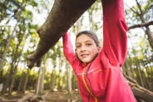Mädchen in pinker Jacke hängt lachend an einem Ast im Gespensterwald Nienhagen, umgeben von hohen Bäumen.