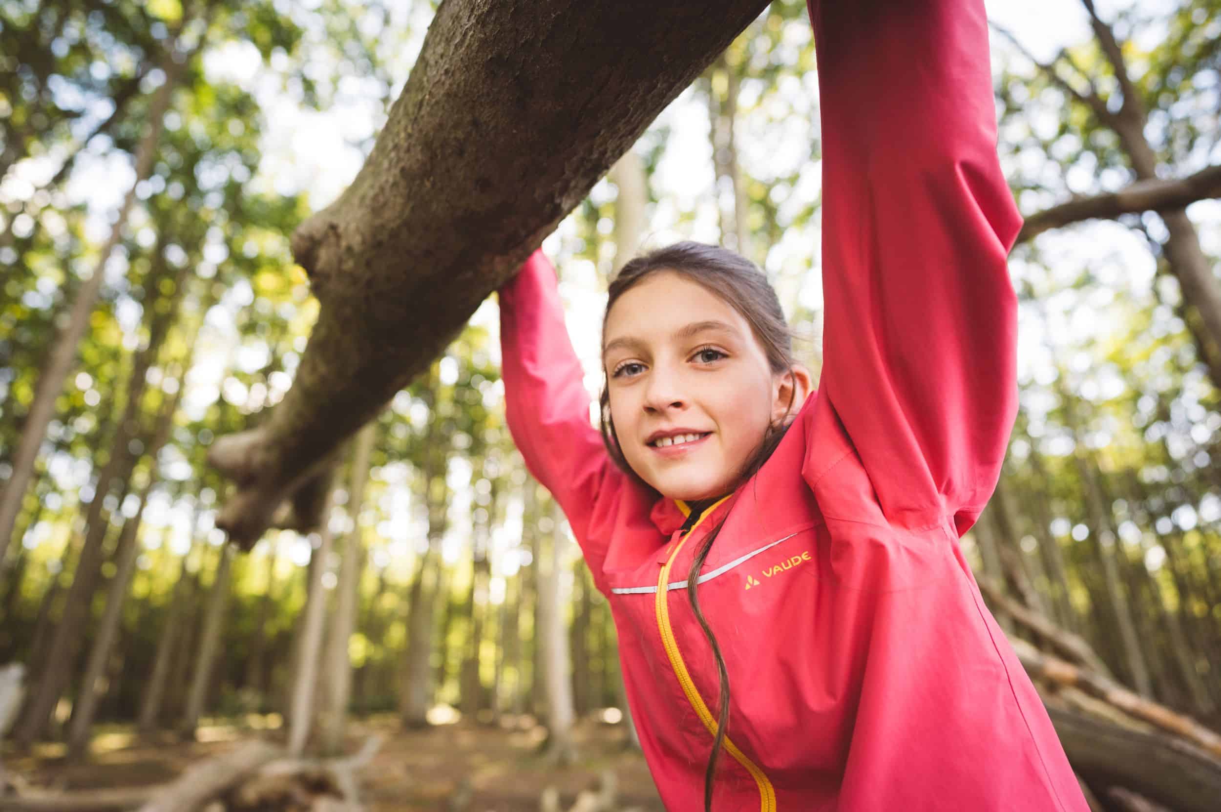 Spiel und Spaß im Gespensterwald Nienhagen – Kletterabenteuer unter Bäumen Mädchen in pinker Jacke hängt lachend an einem Ast im Gespensterwald Nienhagen, umgeben von hohen Bäumen.