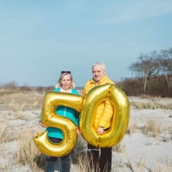 Älteres Paar steht mit goldenen Luftballons in Form der Zahl 50 an einem sonnigen Strand, als Symbol für ein 50-jähriges Jubiläum oder Hochzeitstag.