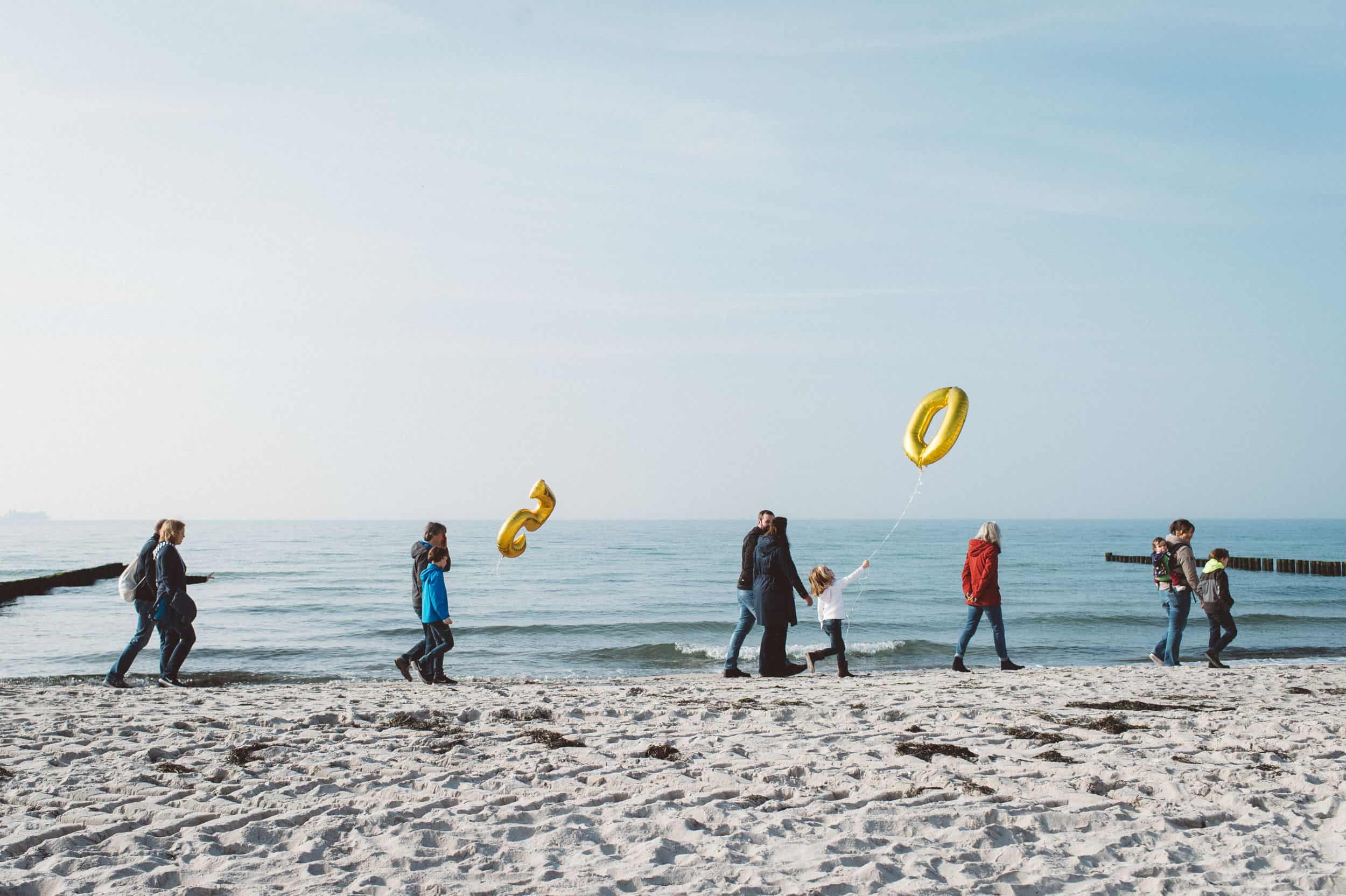 Natürliche Familienfotos am Strand – echte Momente mit Meerblick Mehrere Familien spazieren mit Kindern und goldenen Luftballons in Form von Zahlen am Strand entlang – natürliche Familienfotos mit Ostsee-Flair