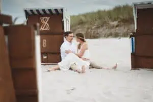 Paar sitzt barfuß in heller Kleidung eng umschlungen im Sand zwischen zwei Strandkörben an der Ostsee in Ahrenshoop.
