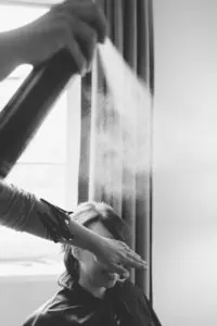 Friseurin sprüht Haarspray auf die Brautfrisur, während die Braut lachend das Gesicht schützt – humorvoller Moment beim Getting Ready.