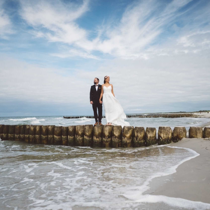 Romantisches Brautpaar am Strand von Ahrenshoop – unvergessliche Hochzeitsmomente vom Hochzeitsfotograf Ahrenshoop Ein Brautpaar steht händchenhaltend auf eine Buhne in Ahrenshoop.