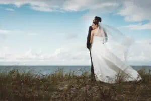 Bräutigam mit Braut in einem Hochzeitskleid und Schleier steht auf einer Düne und blickt auf das Meer und den Himmel in Ahrenshoop.