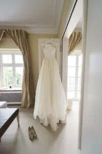 Elegantes, cremefarbenes Brautkleid auf einem Bügel hängend vor einem hellen Fenster im Hotelzimmer mit passenden Hochzeitsschuhen auf dem Boden.